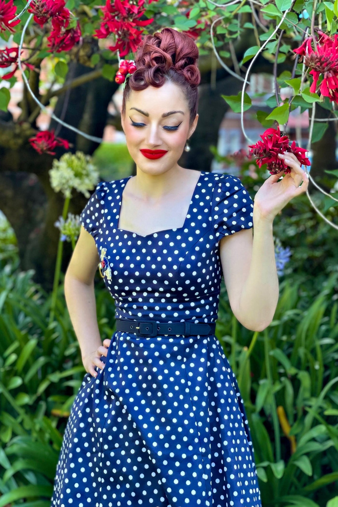 Customer wears our cap sleeved swing dress, in dark blue polka dot print, in front of a flower bush