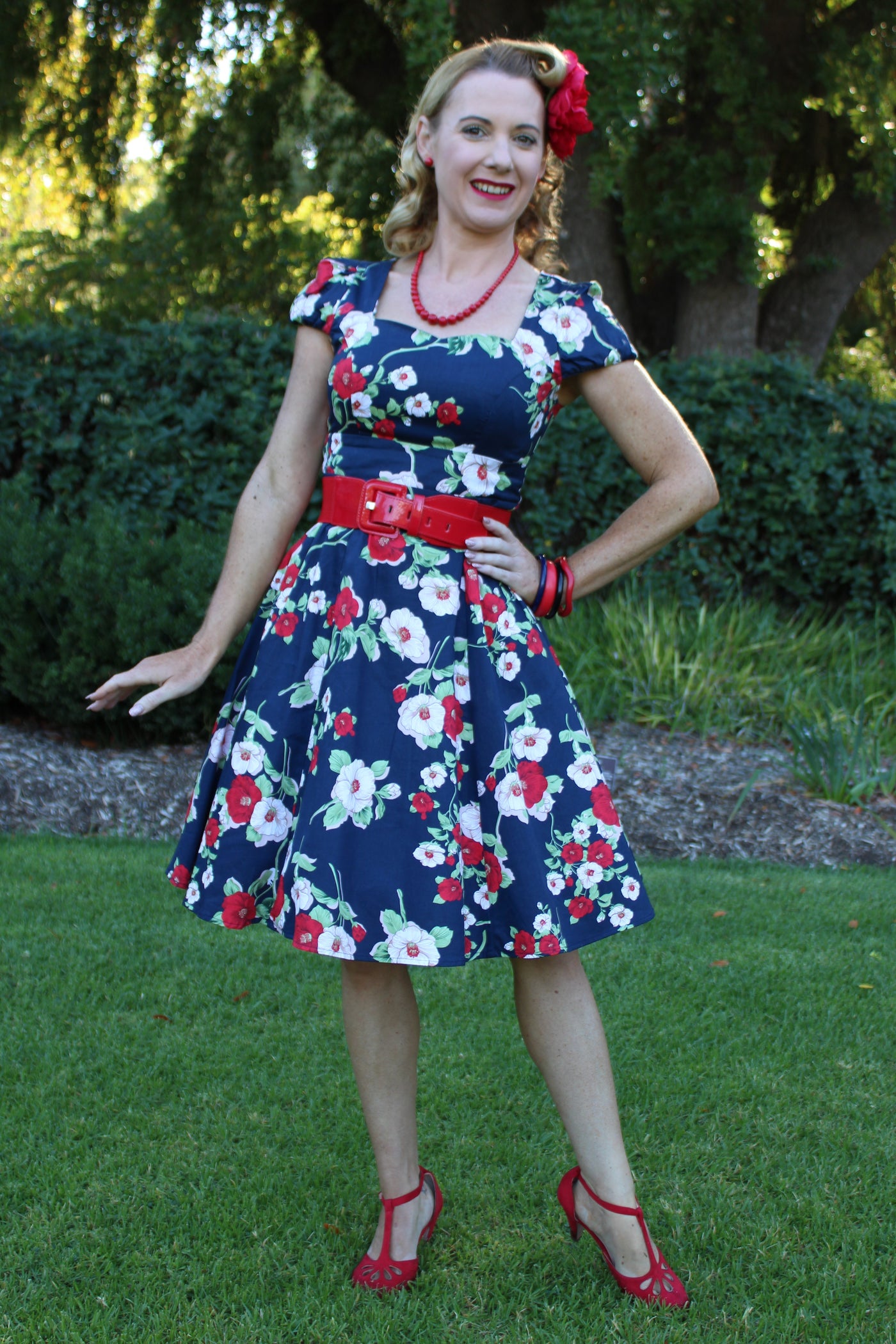 Blogger wears our cap sleeved swing dress, in navy blue, red and white floral print, in front of trees
