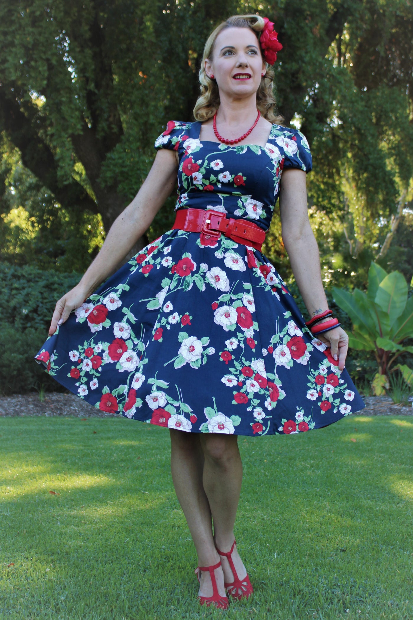 Blogger wears our cap sleeved swing dress, in navy blue, red and white floral print, in front of trees