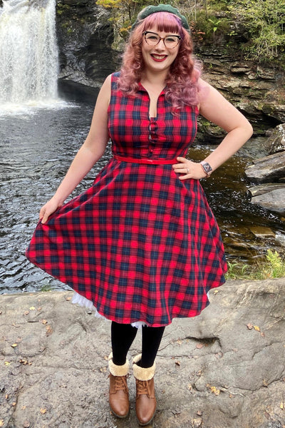 Woman wears our sleeveless Poppy dress, in red and blue tartan print, with accessories, in front of a waterfall and rocks