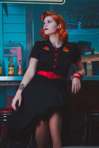 Red headed woman wears our short sleeved Sherry diner dress in black, with red buttons, belt and roses, in a diner