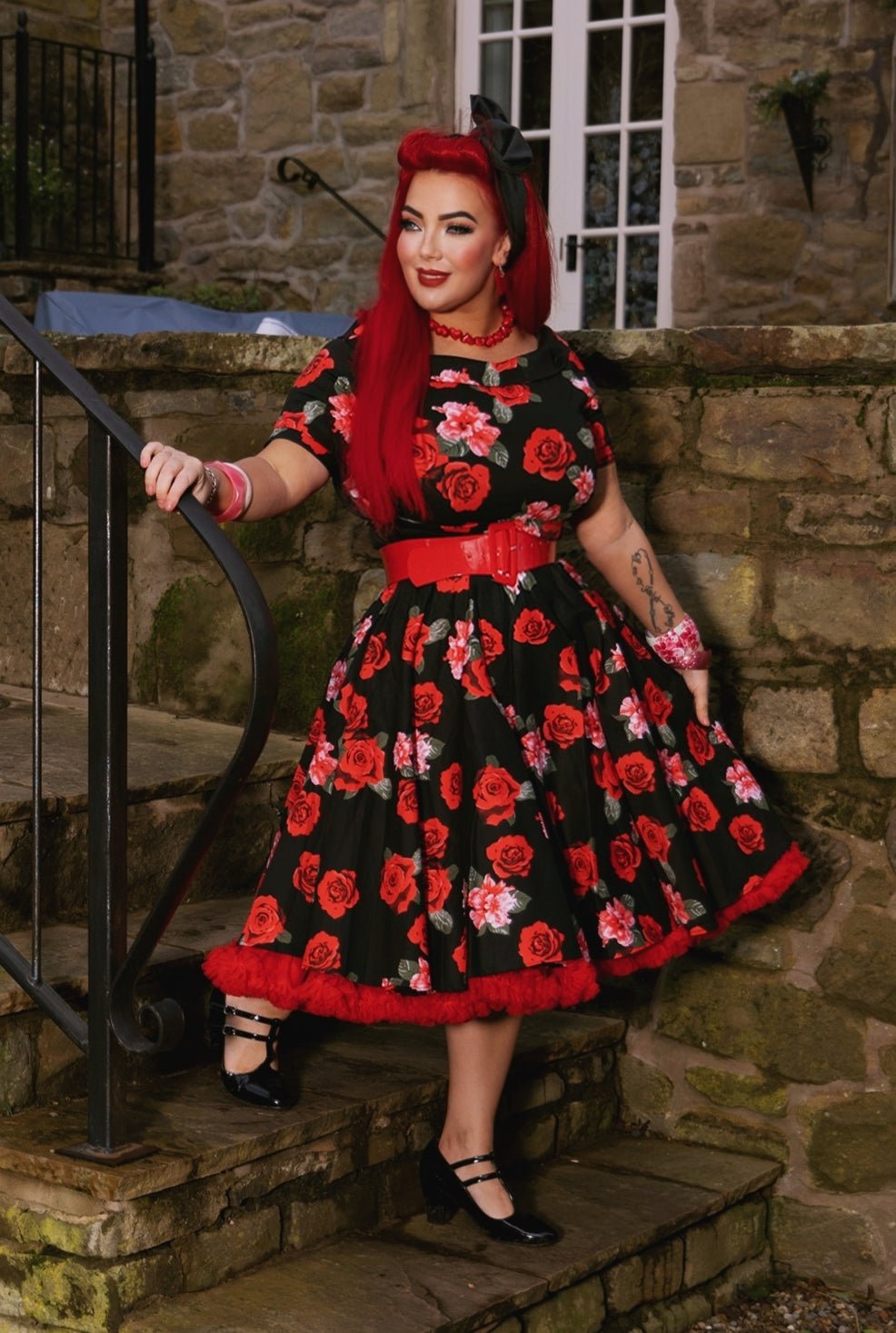 Woman wears our short sleeved swing dress, in black/red roses print, with red accessories, on steps