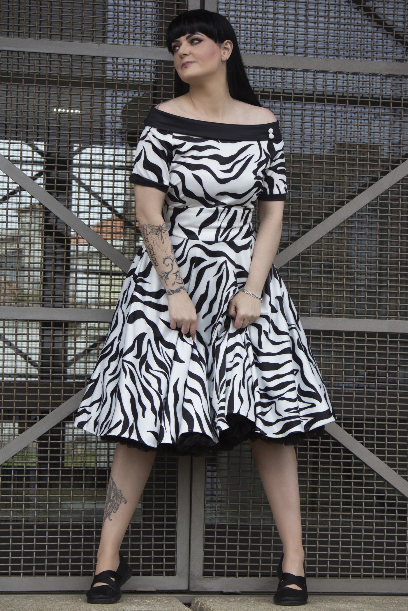 Woman wears our short sleeve flared dress, in white and black zebra print, with a black petticoat, against a metal gate
