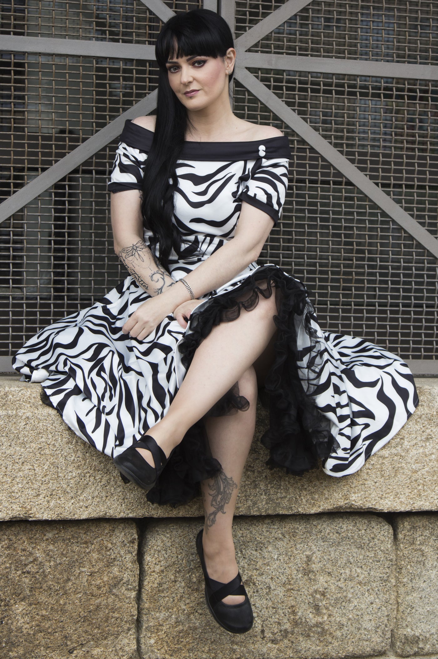 Model wears our short sleeve flared dress, in white and black zebra print, with a black petticoat, sits on a wall