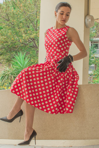 Woman wearing our sleeveless Annie dress, in red, with white spots, with accessories, sat on a wall