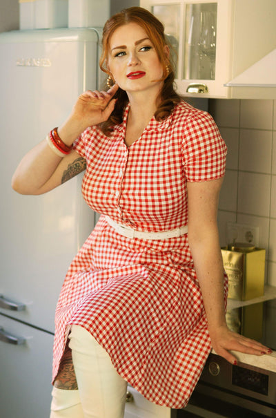Woman wears our short sleeve Penelope dress, in red and white gingham print, with jeans, in a kitchen