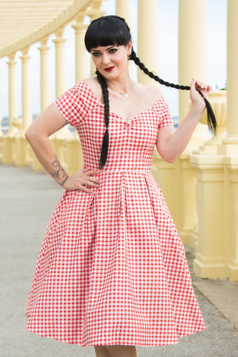 Woman wears our short sleeved, off-shoulder flared dress, in red gingham print, in front of pillars