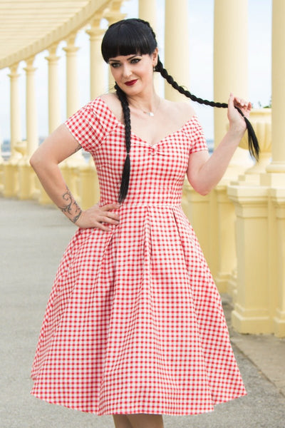 Woman wears our short sleeved, off-shoulder flared dress, in red gingham print, in front of pillars