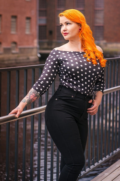 Woman wearing our Gloria off-shoulder Top in Black/White polka dot print, with black jeans, in front of a canal