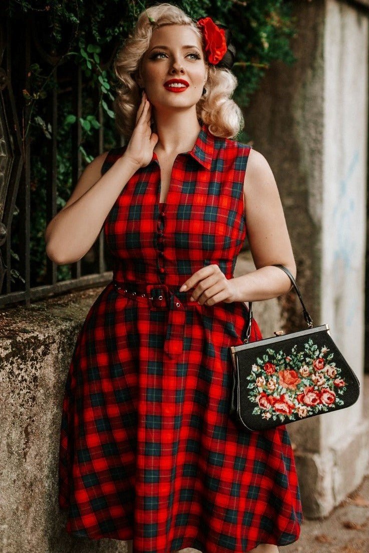 Woman wears our sleeveless Poppy dress, in red and blue tartan print, with accessories, next to a wall and fence