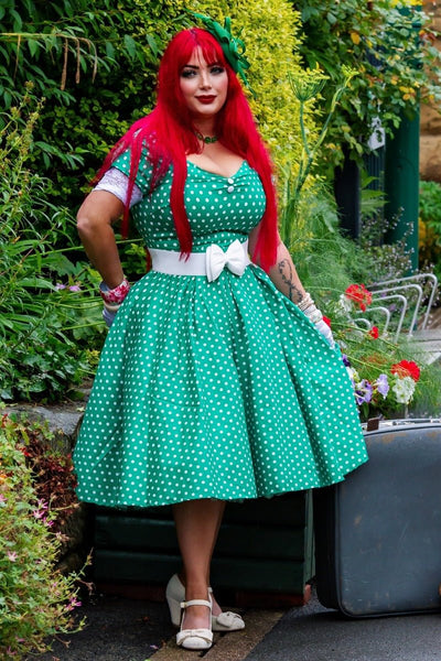 Woman wears our short sleeved, flared dress, in green/white polka dot print, in front of plants