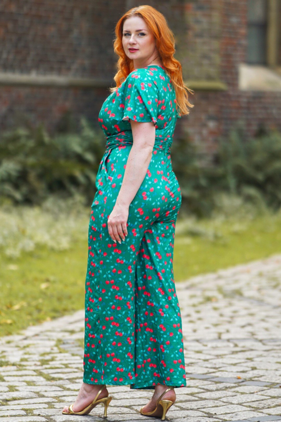Side back view of model wearing our short sleeve jumpsuit in green, with red cherries