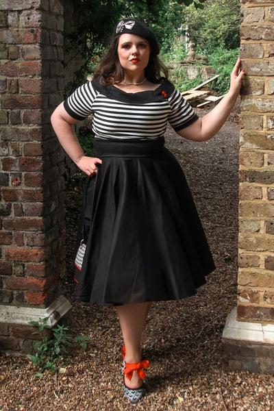 Woman wears our bateau neckline Darlene dress, in black and write stripes, with accessories, next to a brick wall