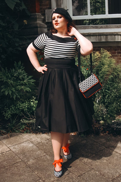 Woman wears our bateau neckline Darlene dress, in black and write stripes, with accessories, in front of a bushes