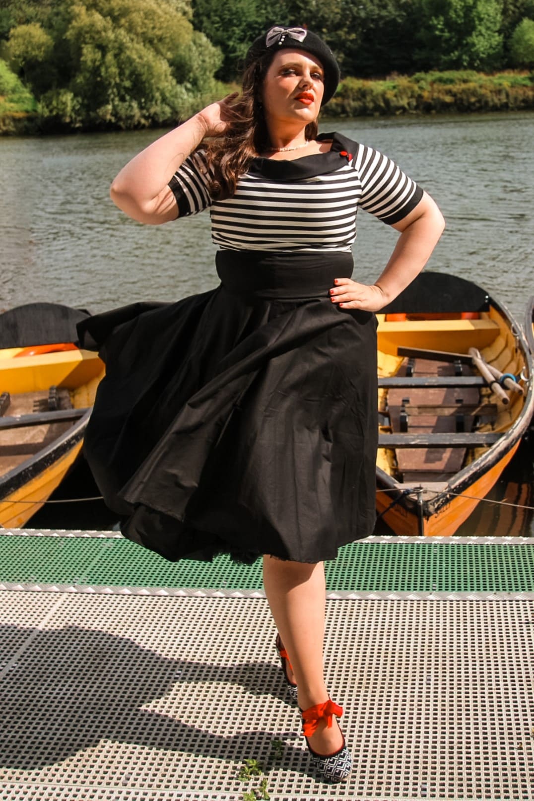 Woman wears our bateau neckline Darlene dress, in black and write stripes, with accessories, in front of a boating lake