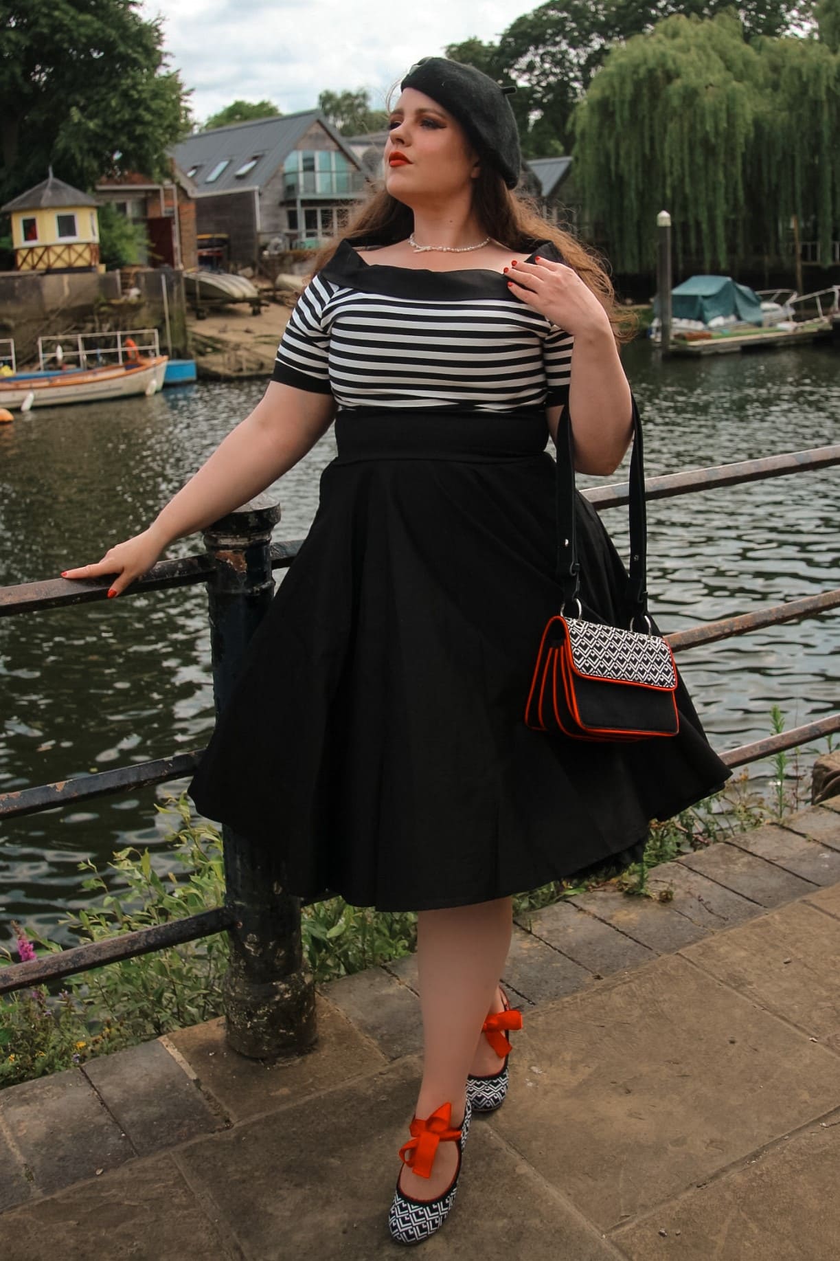 Woman wears bateau neckline Darlene dress, in black and write stripes, in front of a boating lake