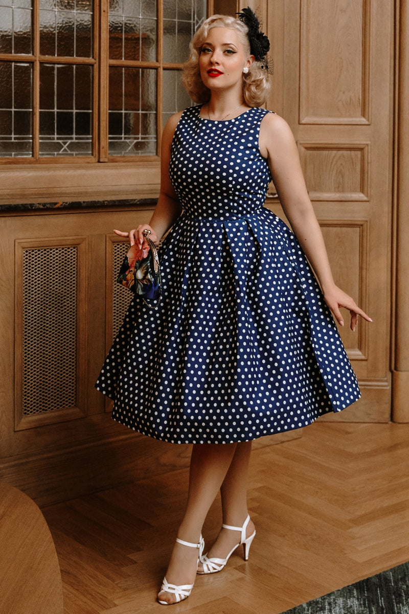 Model wears our sleeveless Lola dress, in dark blue, with white polka dots, with accessories