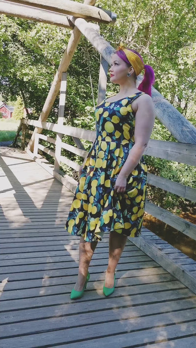 Video of a model on a wooden bridge and a brook wearing our Amanda Vintage Flared Dress in Black/Yellow Lemon Print.