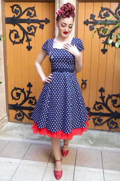 Customer wears our cap sleeved swing dress, in dark blue polka dot print, in front of old gates