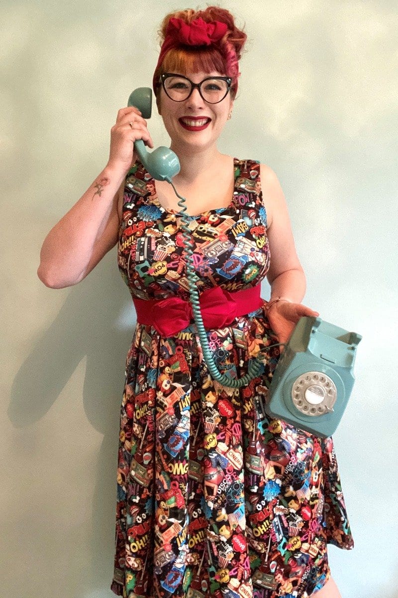 woman wears our Amanda dress, in colourful pop art comic print, whilst holding a vintage telephone
