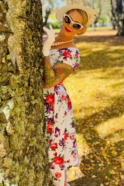Woman wearing our Lily, off-shoulder 50's Evening Dress in White, with pink and purple flowers, whilst hiding behind a tree