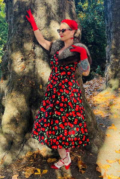 Woman wears our V neck midi flared dress, in black, red cherry print, with red accessories, in front of a tree
