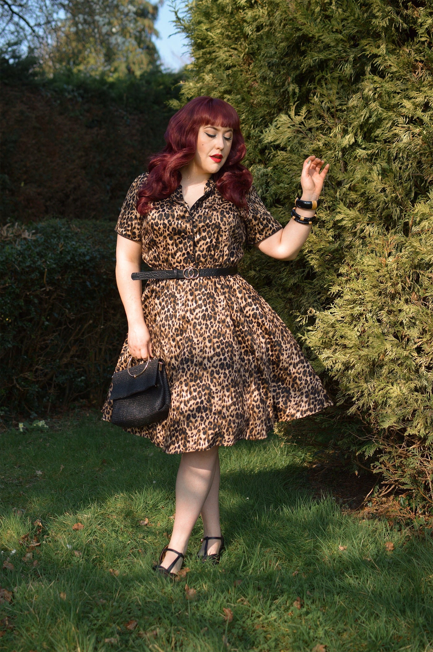 Woman wears our short sleeve Penelope dress, in brown leopard print, in a park, with hedges