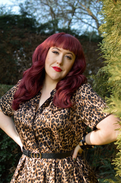 Woman wears our short sleeve Penelope dress, in brown leopard print, in a park, with hedges
