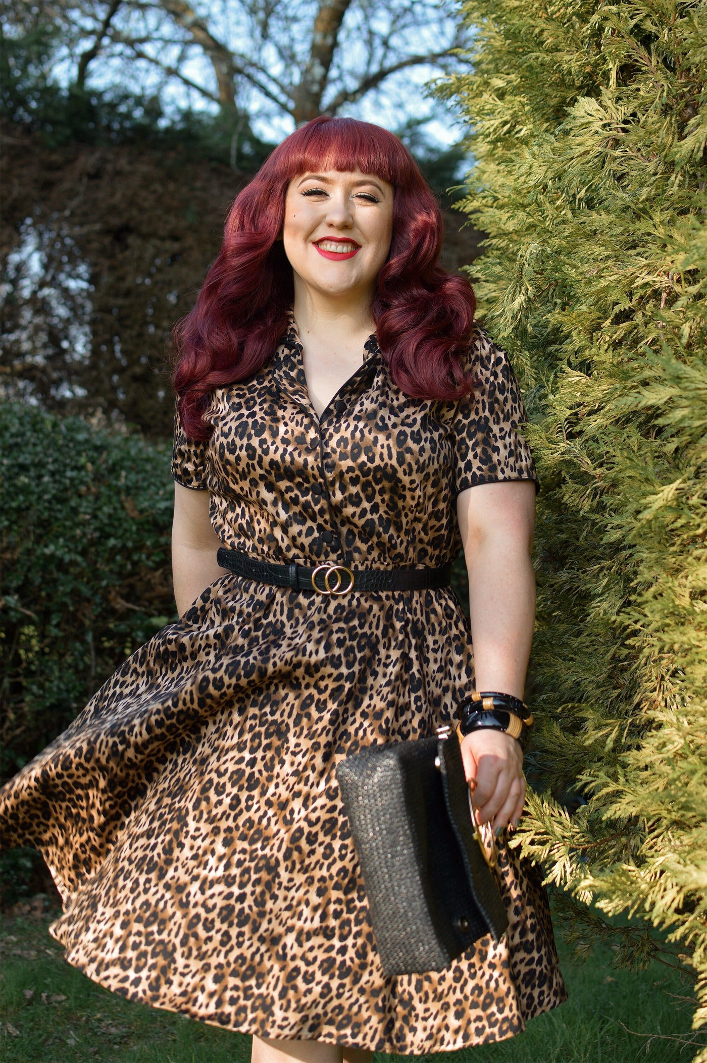 Woman wears our short sleeve Penelope dress, in brown leopard print, in a park, with hedges