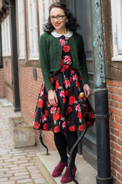Woman wearing our Annie sleeveless swing dress, in black, with red and pink roses, with green cardigan, next to a building