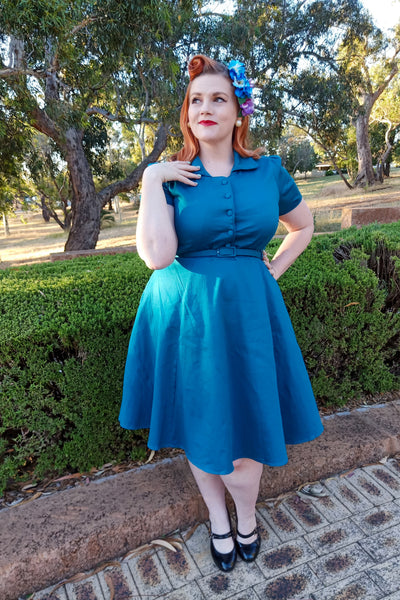 Woman wearing our short sleeved Penelope button top dress in teal blue, in front of trees