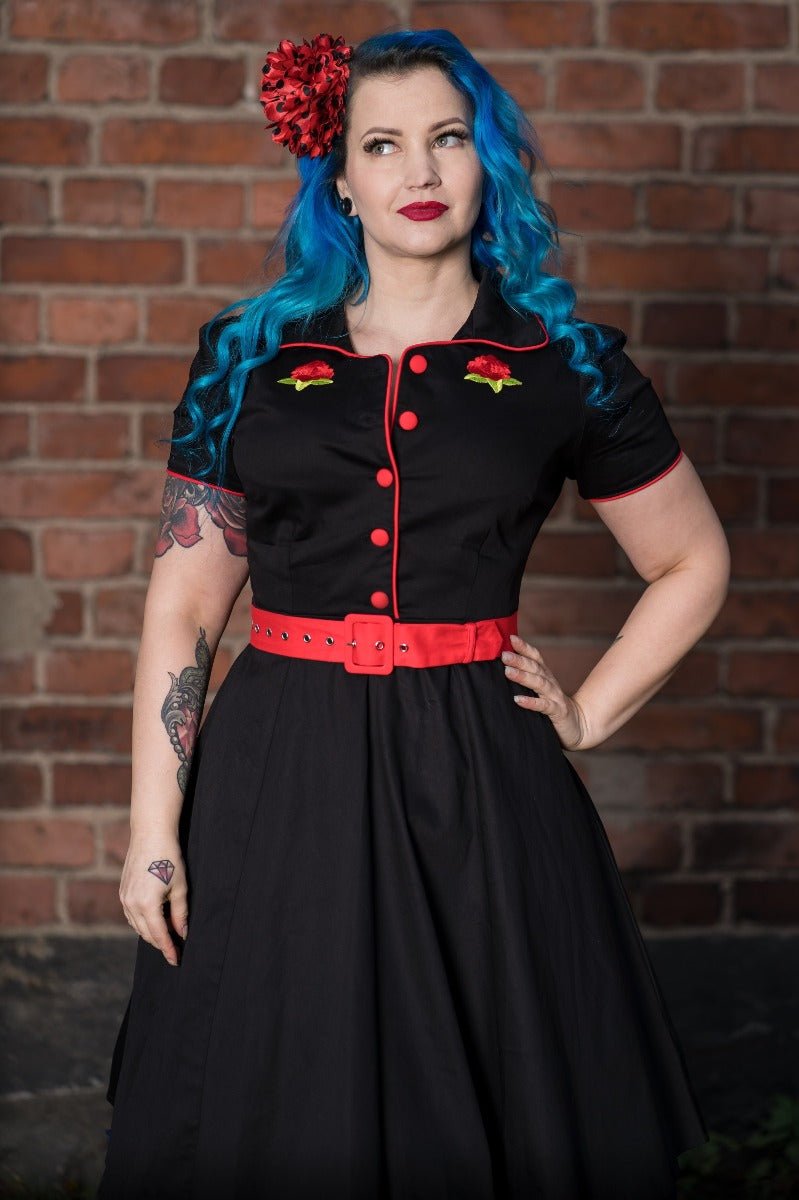 Woman with tattoos, wears our short sleeved Sherry diner dress in black, with red buttons, belt and roses, in front of a brick wall