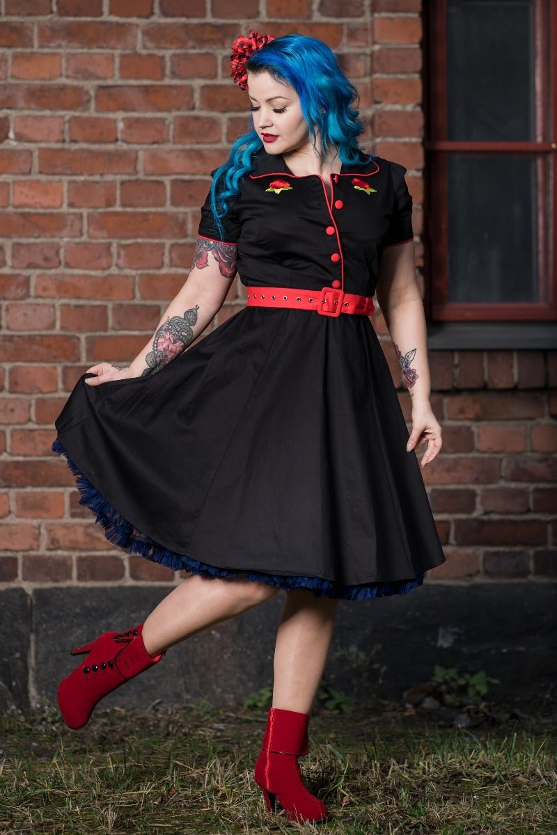 Woman wears our short sleeved Sherry diner dress in black, with red buttons, belt and roses, in front of a brick wall