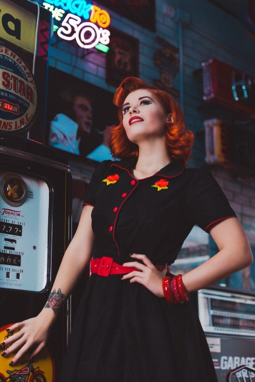 model wears our short sleeved Sherry diner dress in black, with red buttons, belt and roses, in an American diner