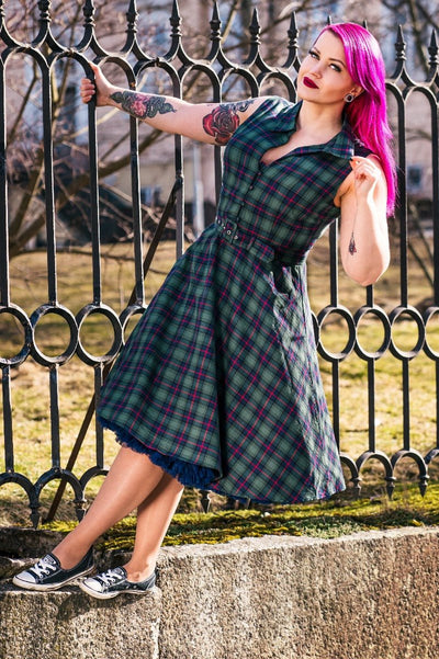 Pink haired woman, with tattoos wears our Poppy dress in green tartan print, with a petticoat, whilst hanging on a fence