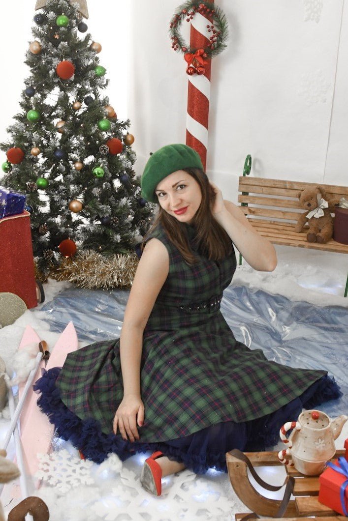 Woman wears our sleeveless Poppy button up dress in green tartan print, with a petticoat and accessories, in front of a christmas tree