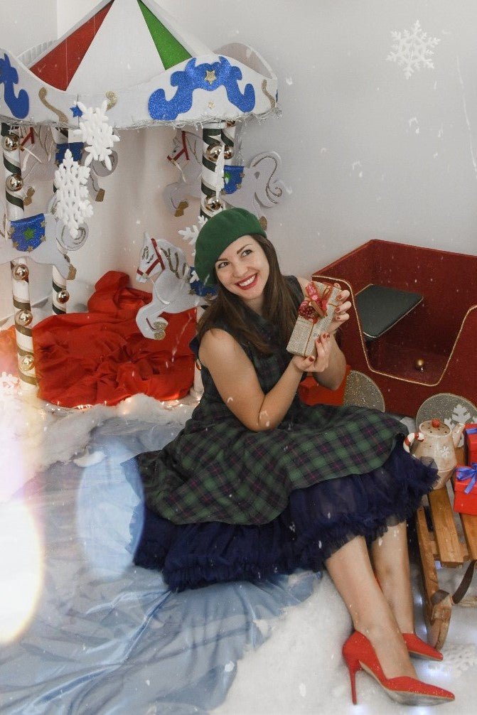 Woman wears our sleeveless Poppy button up dress in green tartan print, with a petticoat and accessories, in a winter wonderland