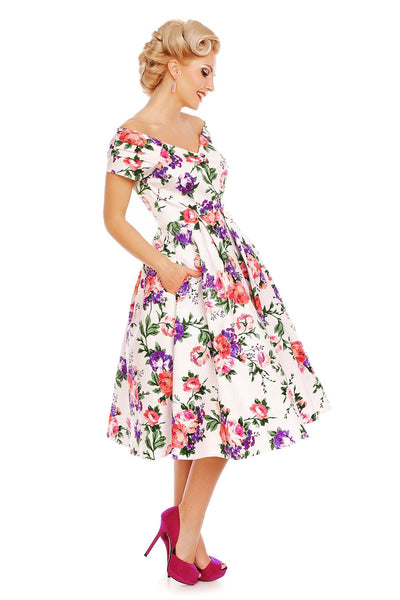 Model wearing our Lily, off-shoulder 50's Evening Dress in White, with pink and purple flowers, side view, hands in her pockets