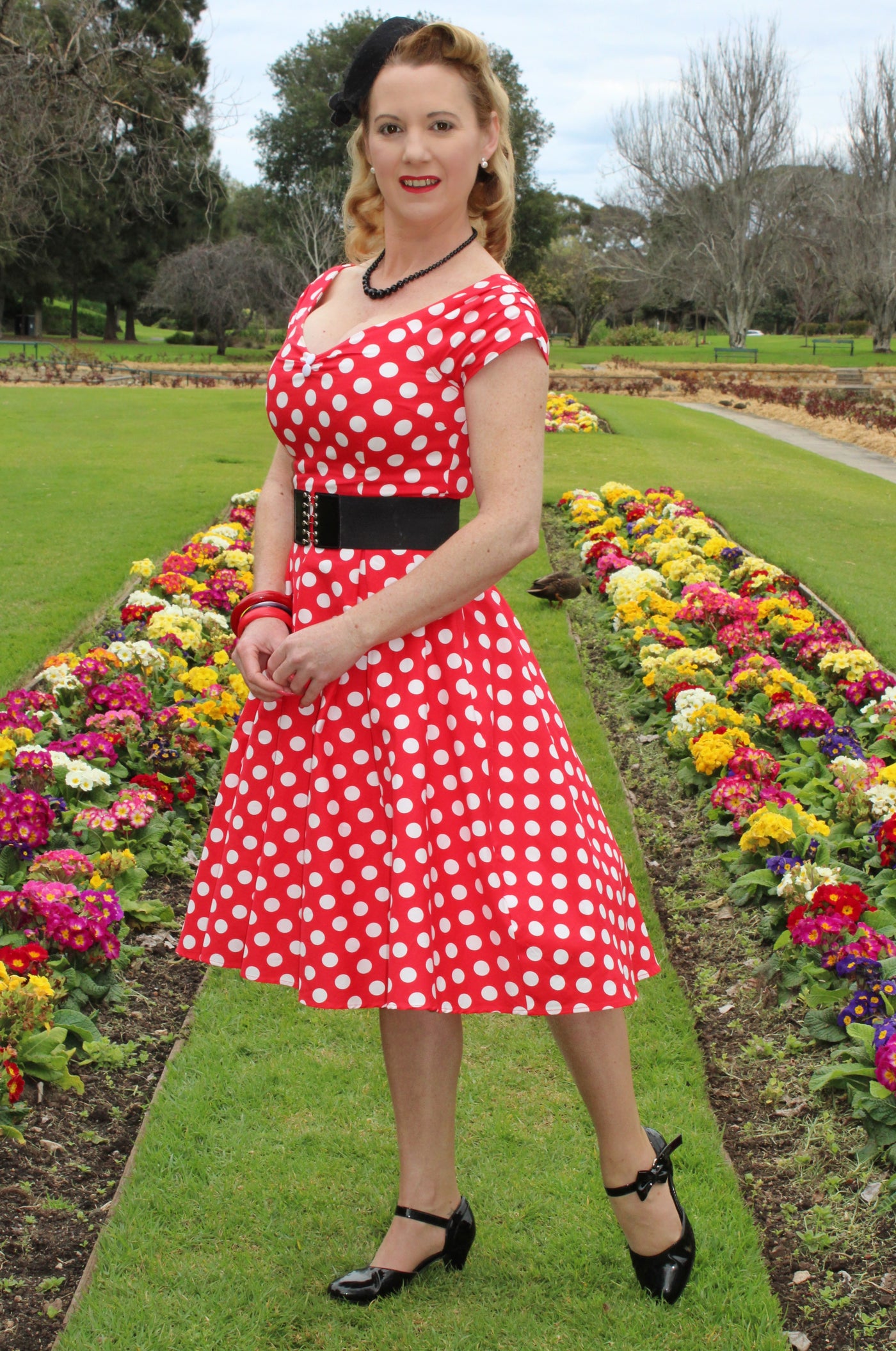 Woman wears our V neck, off-shoulder flared dress, in red, with white polka dots, front view, with flowers