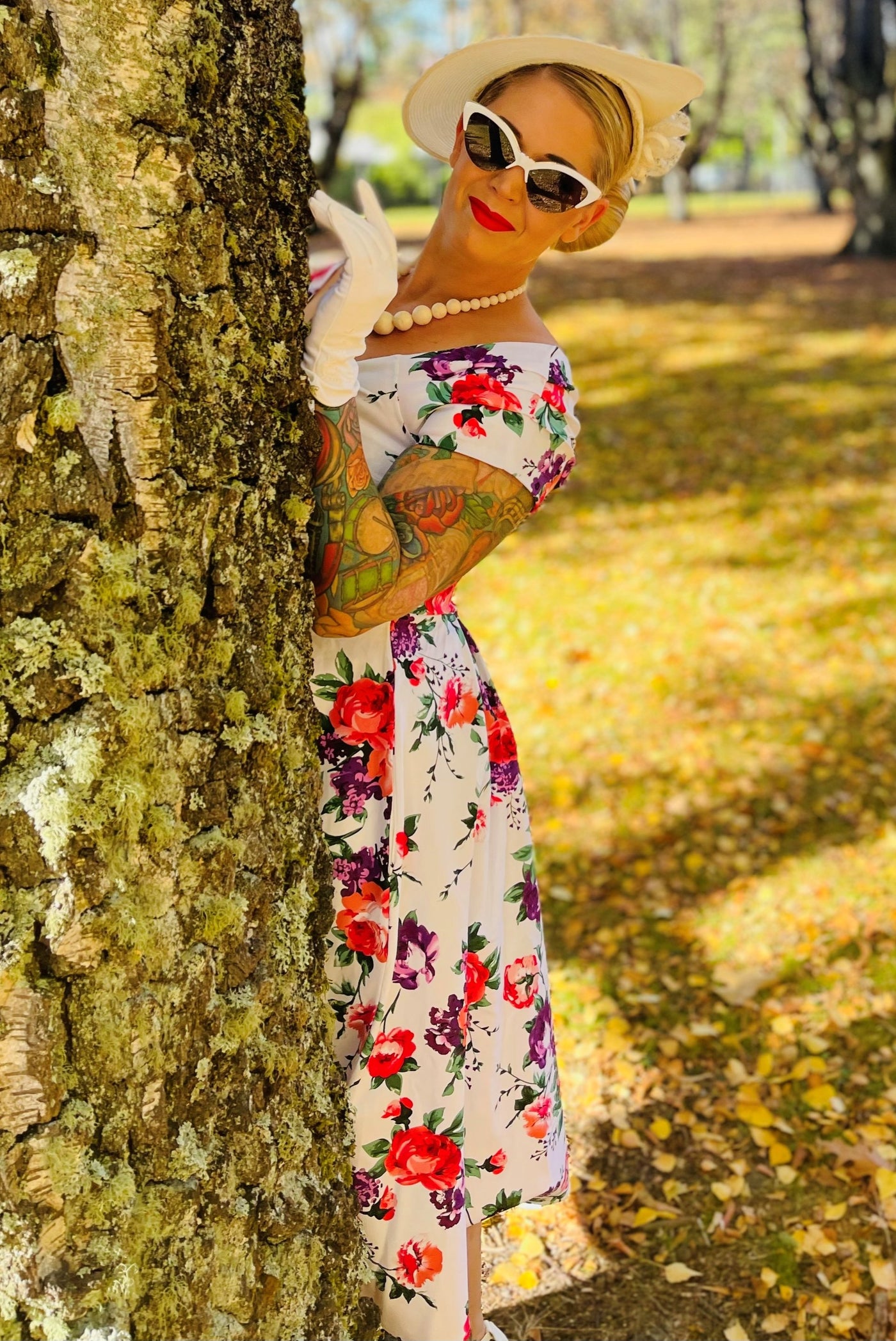 Woman wearing our Lily, off-shoulder 50's Evening Dress in White, with pink and purple flowers, whilst hiding behind a tree
