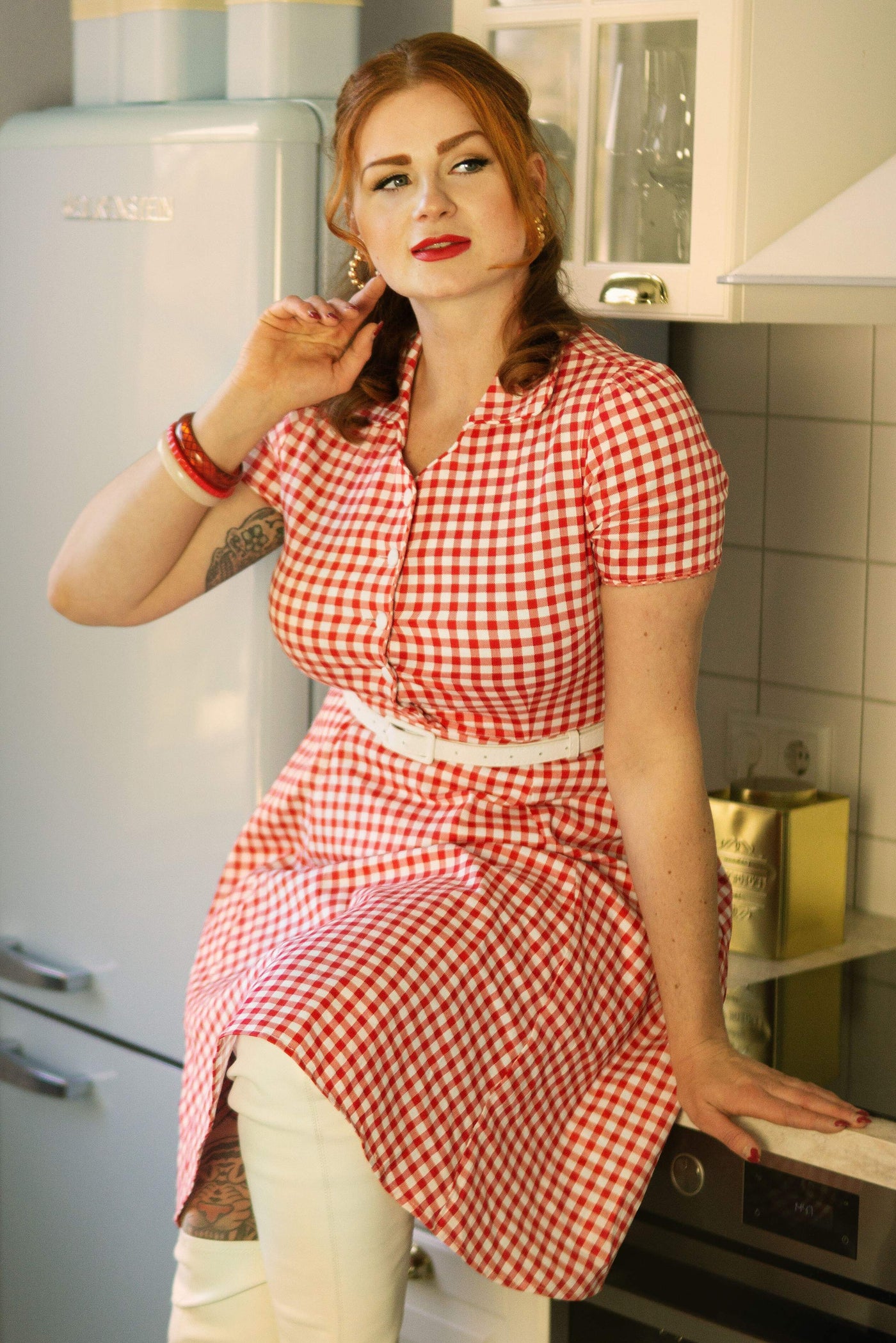 Woman wears our short sleeve Penelope dress, in red and white gingham print, with jeans, in a kitchen