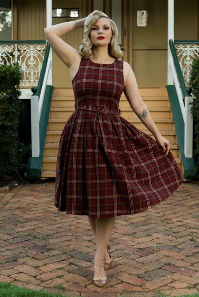 Woman wears our sleeveless Annie swing dress, in burgundy red tartan print, in front of a house
