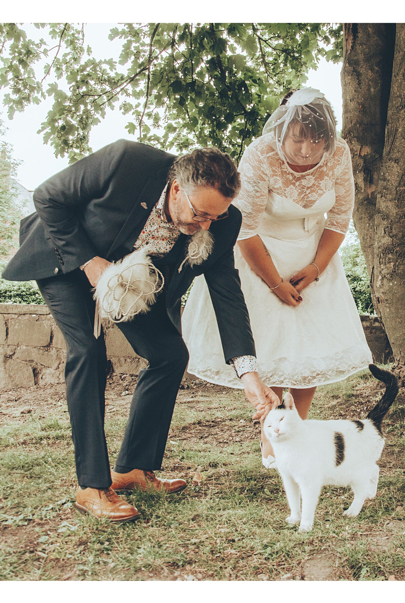 Customer wears our lace covered, sleeved vintage-style wedding dress, with her suited husband, stroking a cat