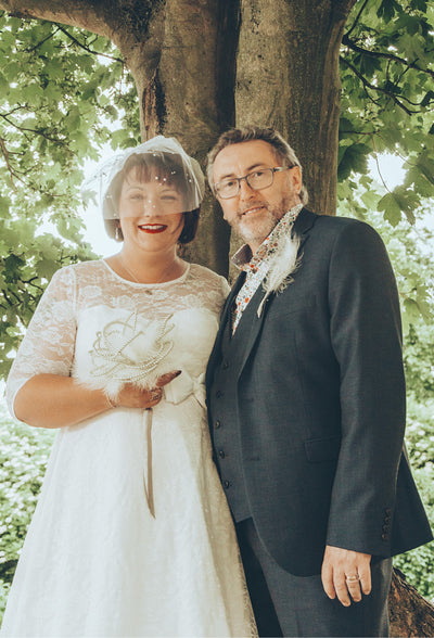 Customer wears our lace covered, sleeved vintage-style wedding dress, with her suited husband, in front of a tree
