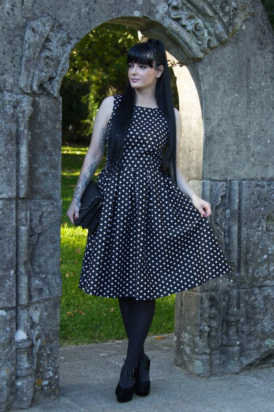 Woman wears our sleeveless Lola dress in black, with white polka dots, with accessories, under an arch