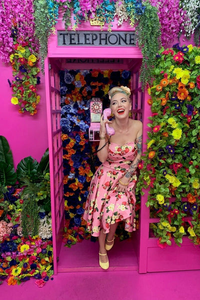 Model wearing strapless swing dress in pink strawberry print in a pink telephone box