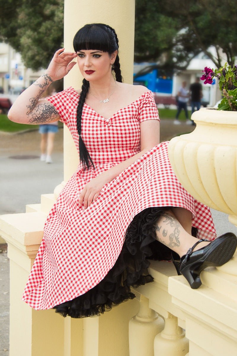 Woman wears our short sleeved, off-shoulder flared dress, in red gingham print, sitting on a stone wall