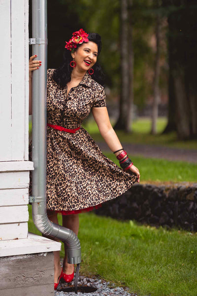 Woman wears our short sleeve Penelope dress, in brown leopard print, whilst holding a drainpipe