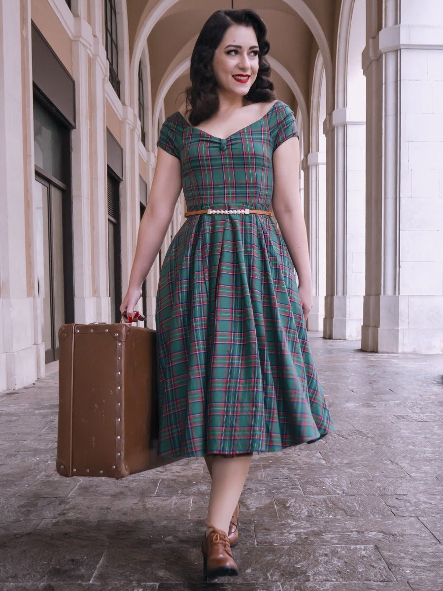 Woman wearing our short sleeved Lily dress in green and red tartan check print, with a suitcase, in an archway