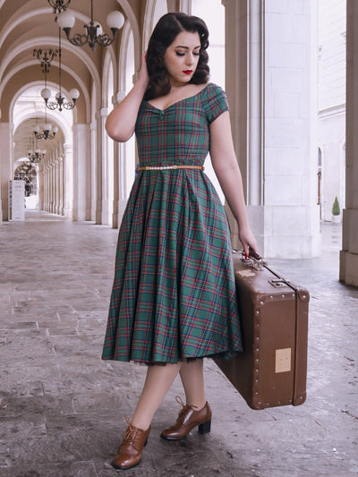 Woman wearing our short sleeved Lily dress in green and red tartan check print, with a suitcase, in an archway